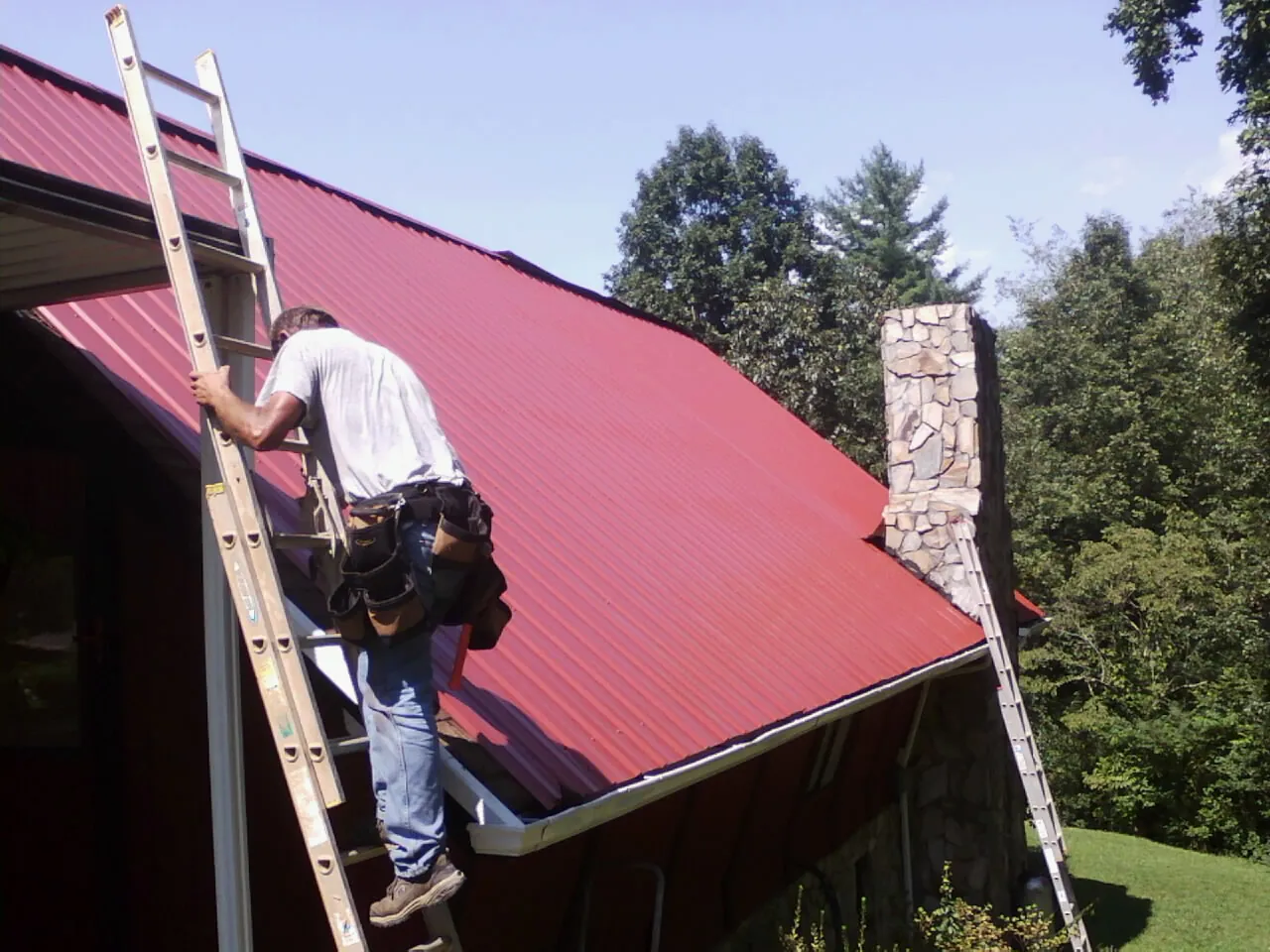 Roofer on a ladder beside a red metal roof and stone chimney — Soffit & Fascia Repair by Roofing Brothers in Hyrum